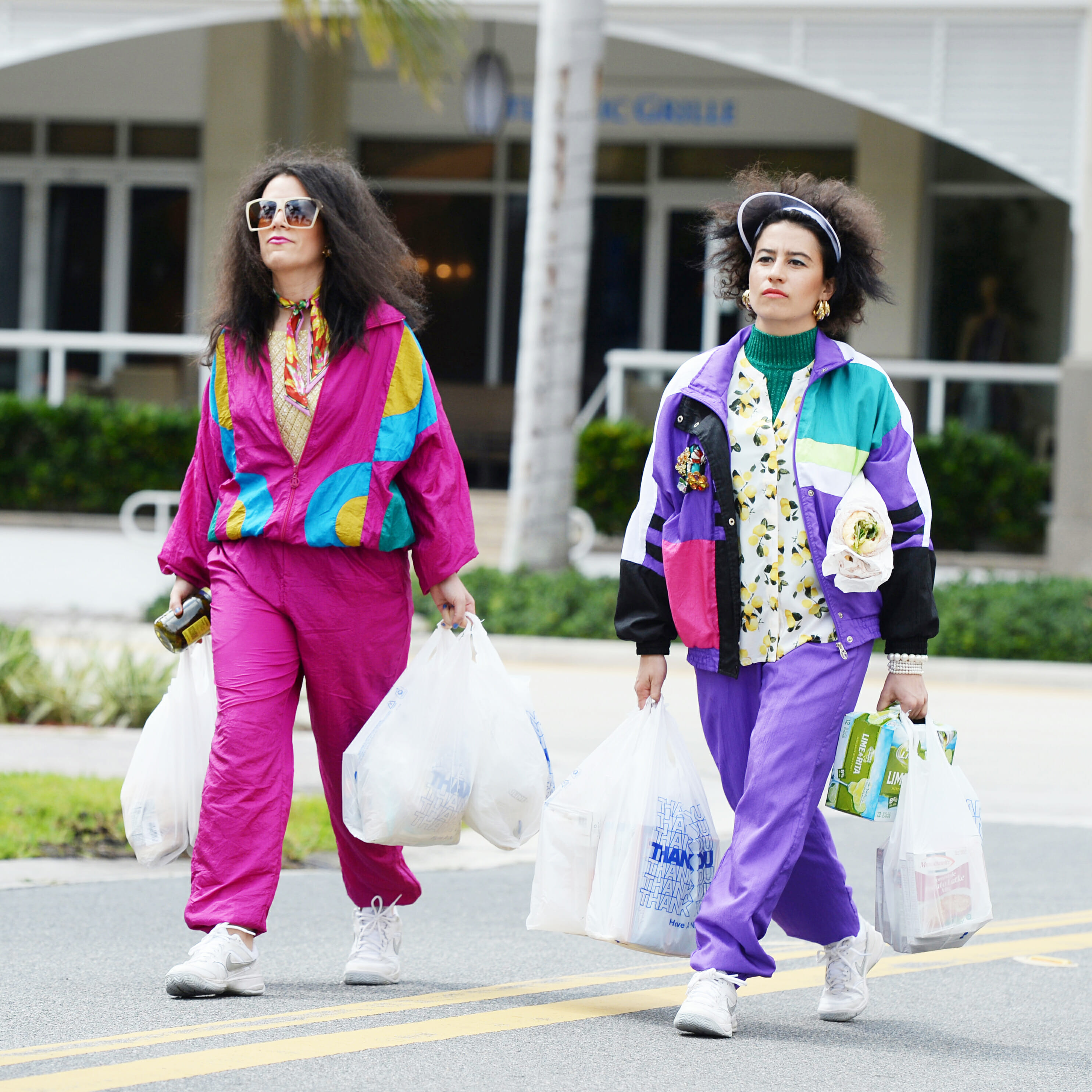 Two women dressed in colorful clothing march out of grocery store carrying many shopping bags in both their hands.