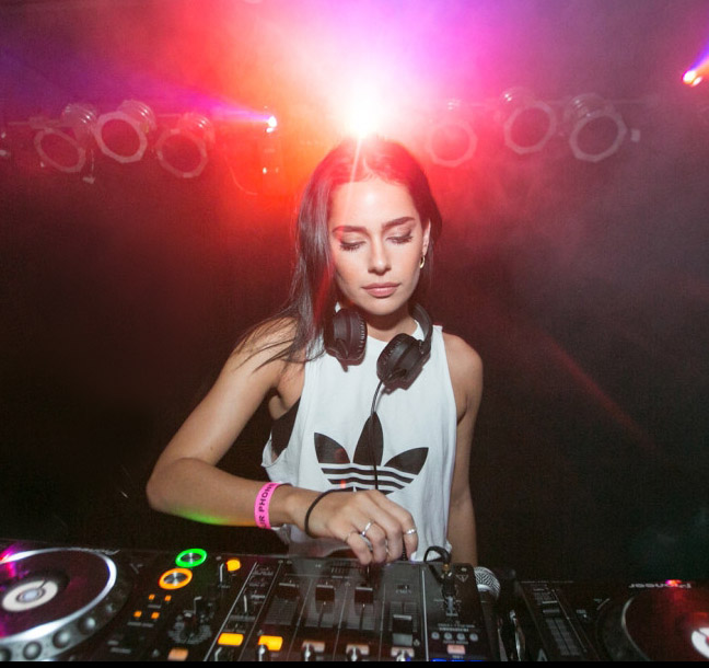 Woman DJ at the set, blarring lights in the background. Woman is wearing sleeveless T, pink bracelet. She's looking down. She has dark hair.